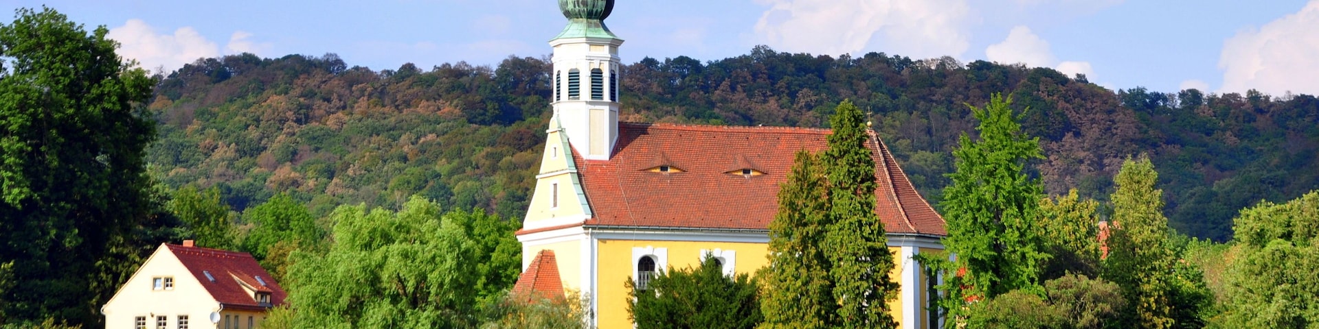Eine Kirche mit einer grünen Kuppel und einem roten Dach, umgeben von Bäumen und einer Hügelkulisse im Hintergrund.