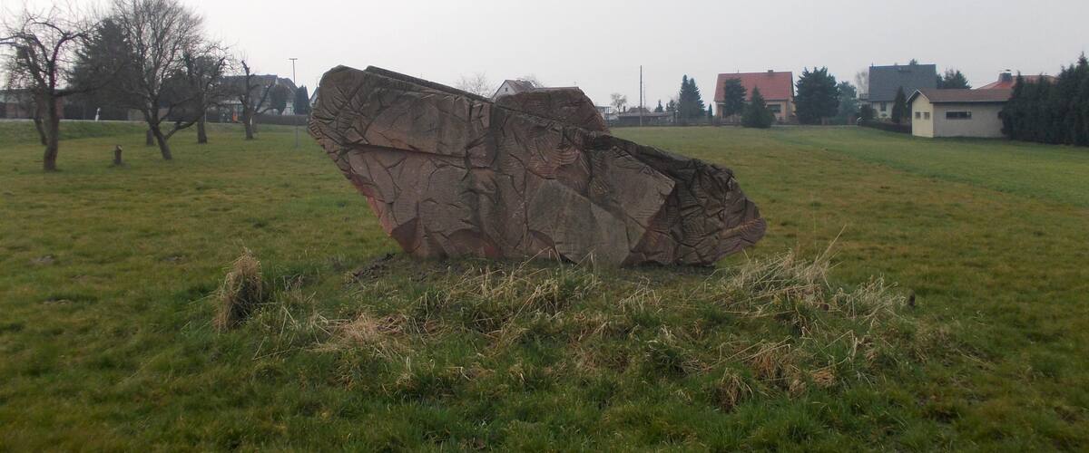 A large rock formation in a grassy field with trees and houses in the background.
