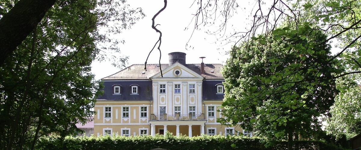 A large, historic building with a symmetrical facade and a central entrance, surrounded by lush greenery and a manicured lawn.