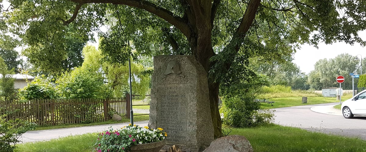 A gravestone with inscriptions, surrounded by grass and flowers, under a tree.