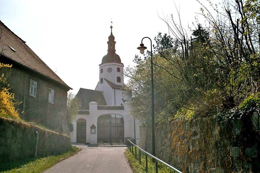 Eine schmale Straße führt zu einer Kirche mit einem hohen Glockenturm und einer Uhr.