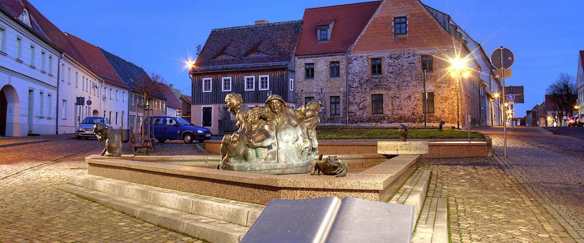 A cobblestone square with a bronze statue in the center, surrounded by historic buildings with red roofs.