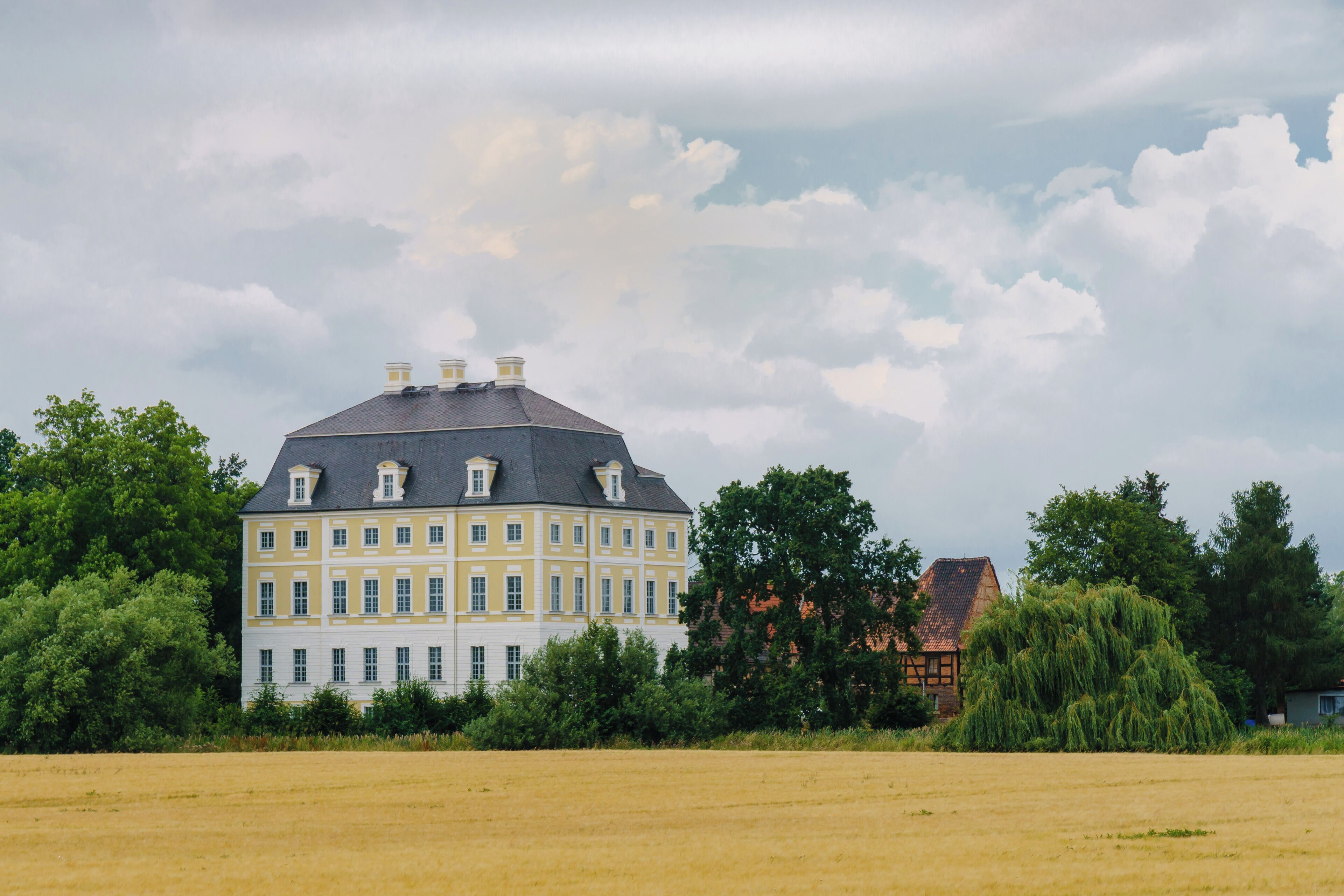 A large yellow building with a grey roof, surrounded by trees and a field.