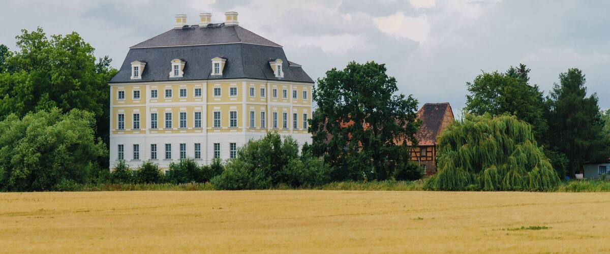 A large yellow building with a grey roof, surrounded by trees and a field.