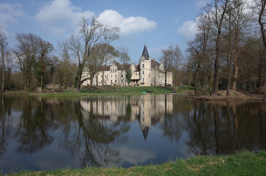 A large, historic building with a tower, surrounded by trees and a calm lake.