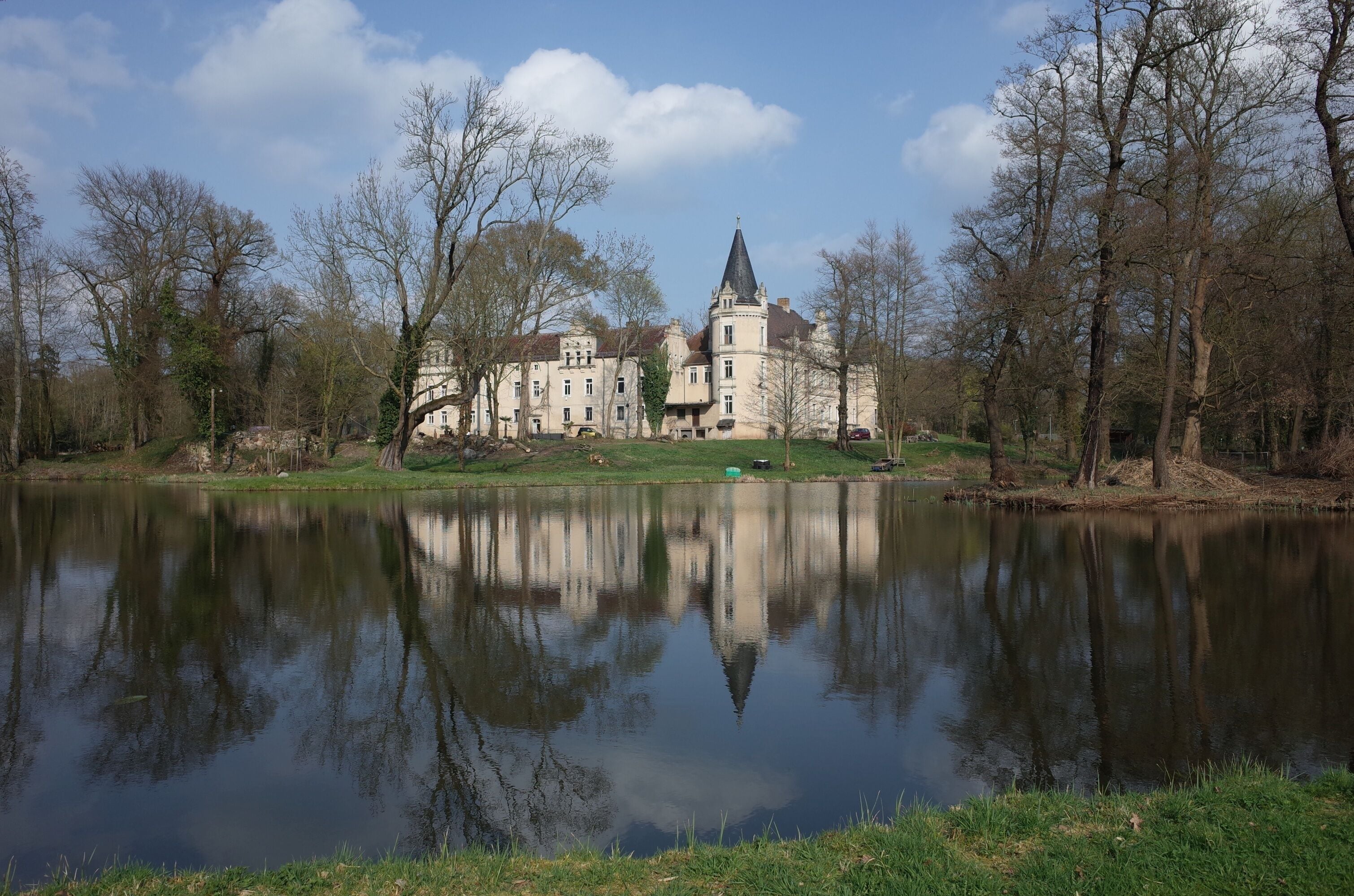 A large, historic building with a tower, surrounded by trees and a calm lake.