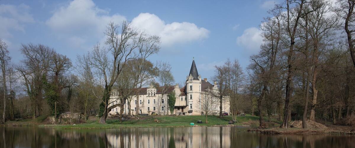 A large, historic building with a tower, surrounded by trees and a calm lake.