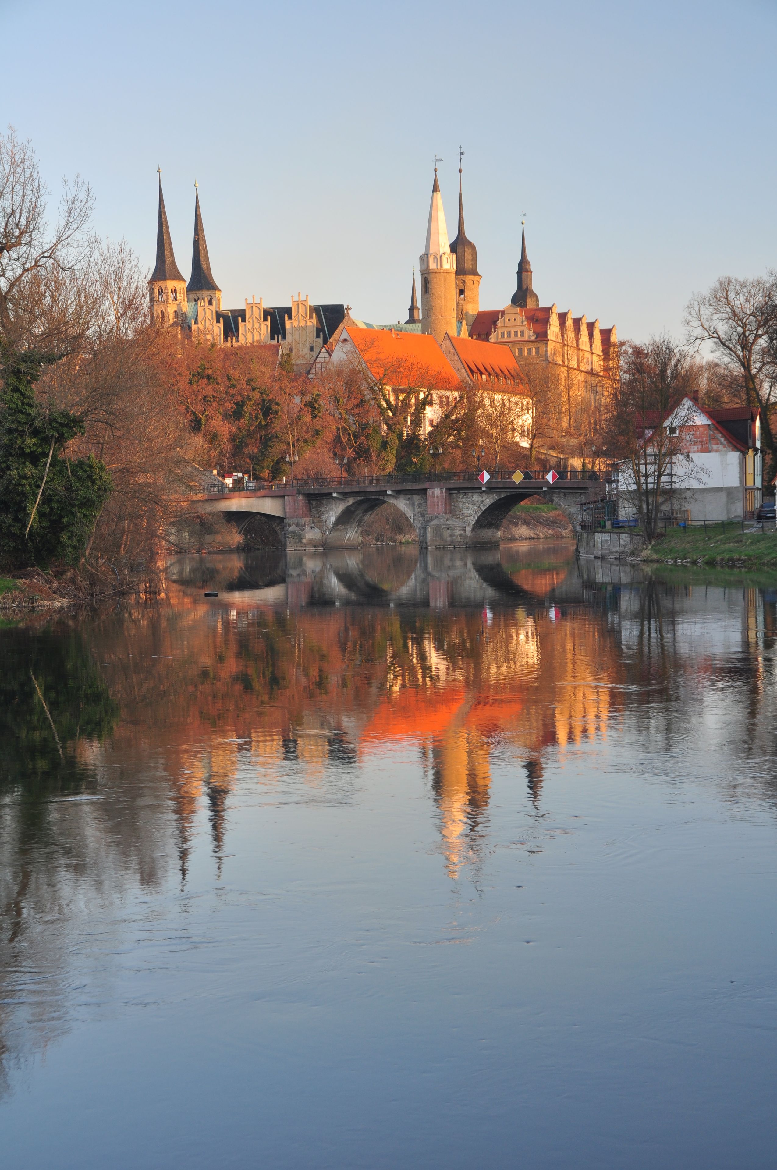A historic castle with multiple spires, a bridge over a calm river, and autumn-colored trees.