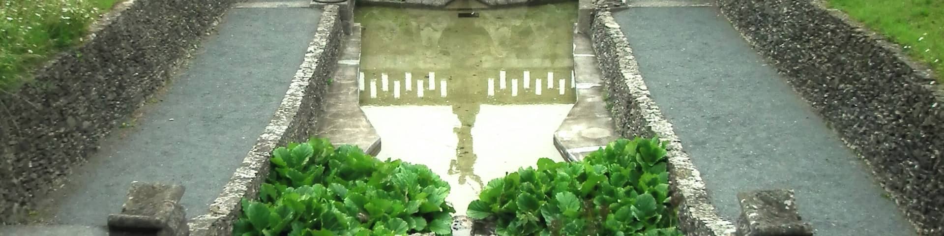 A formal garden with a central water feature, stone statues, and a bridge in the background.