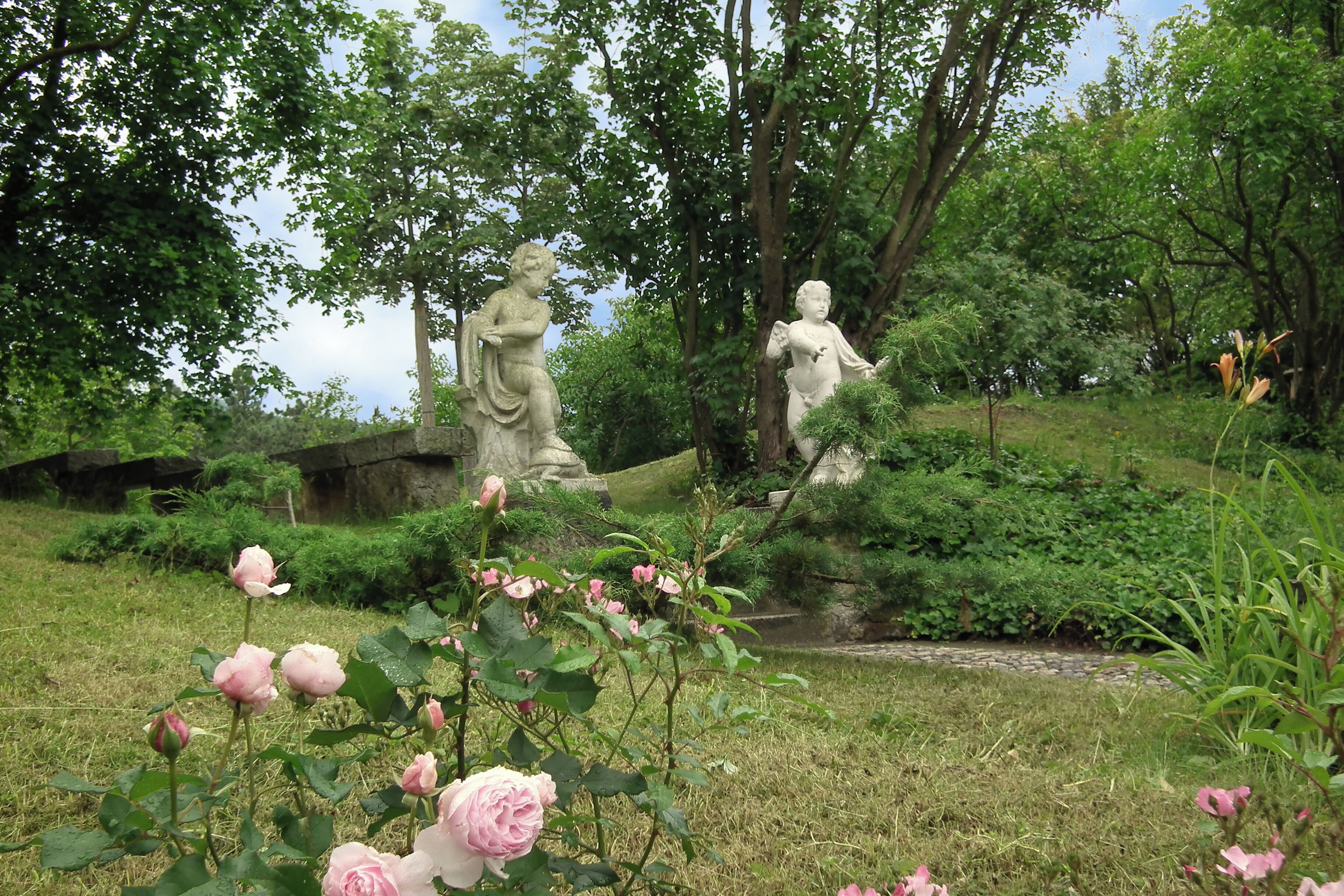 A garden with statues, pink roses, and lush greenery.