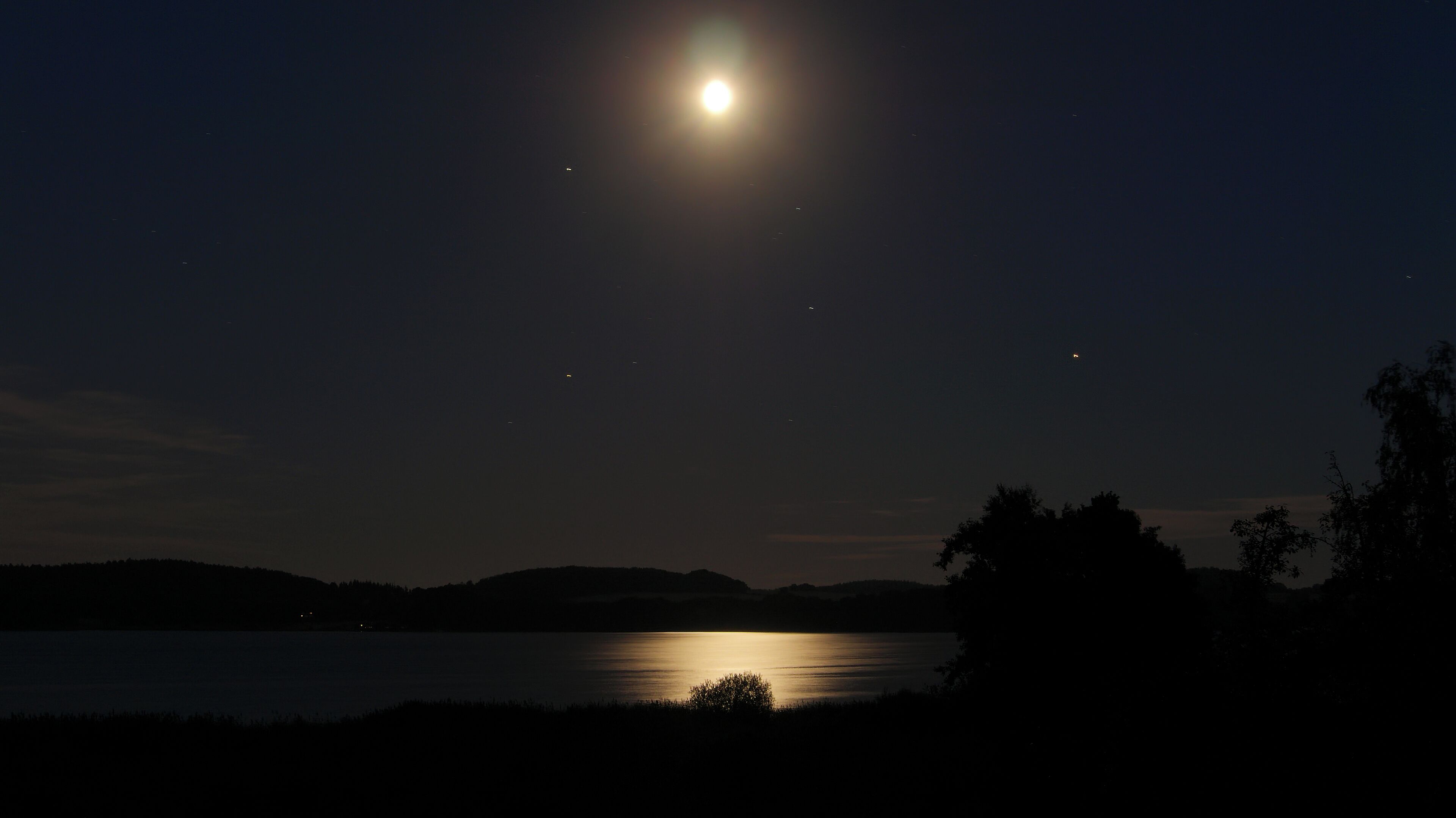 A night scene with a bright moon over a calm lake, surrounded by hills and silhouetted trees.
