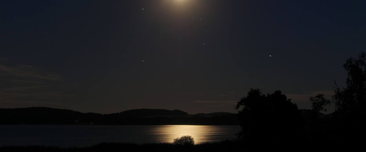 A night scene with a bright moon over a calm lake, surrounded by hills and silhouetted trees.