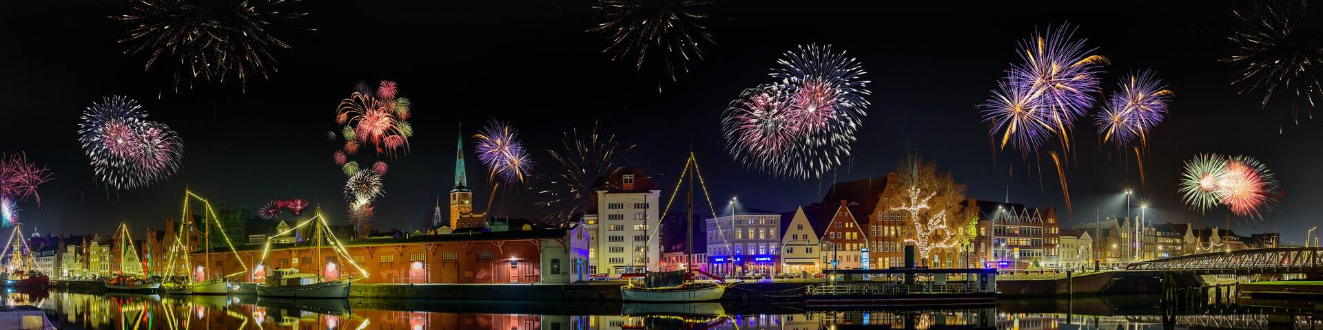 A nighttime cityscape with fireworks over a calm river, illuminated buildings, and a bridge.