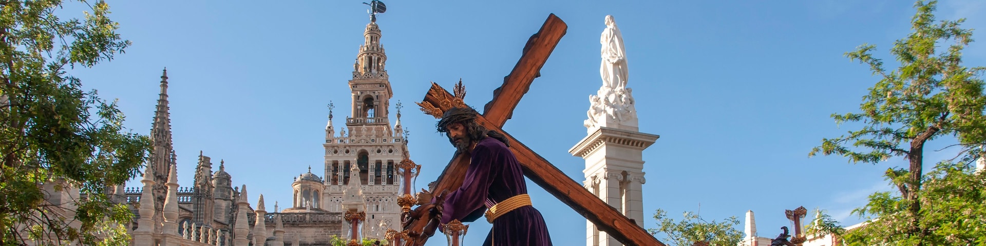 Procissão religiosa com uma figura carregando uma cruz, rodeada por decorações elaboradas e arquitetura histórica ao fundo.