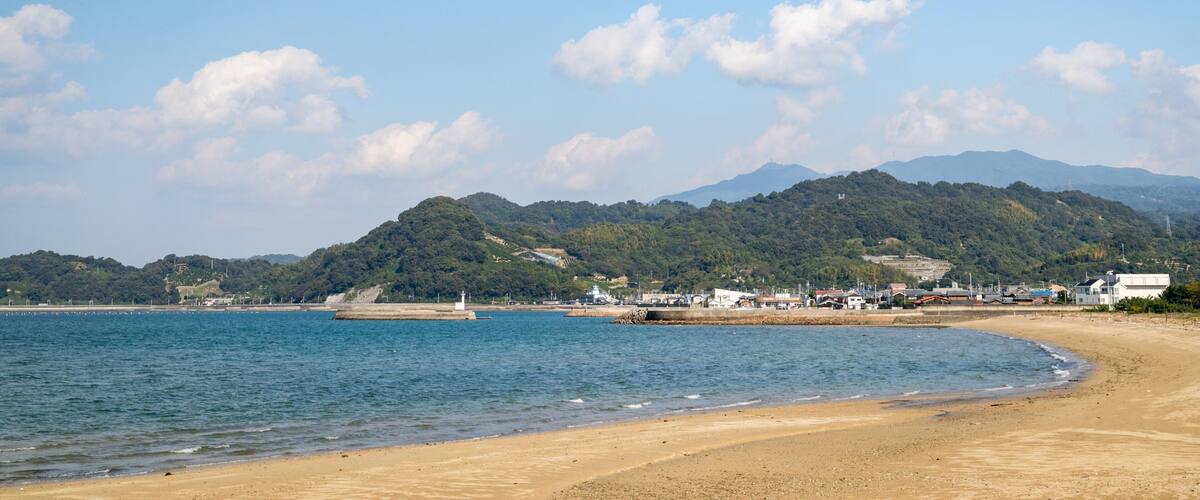 A beach with a sandy shore, calm water, and a backdrop of green hills and mountains.