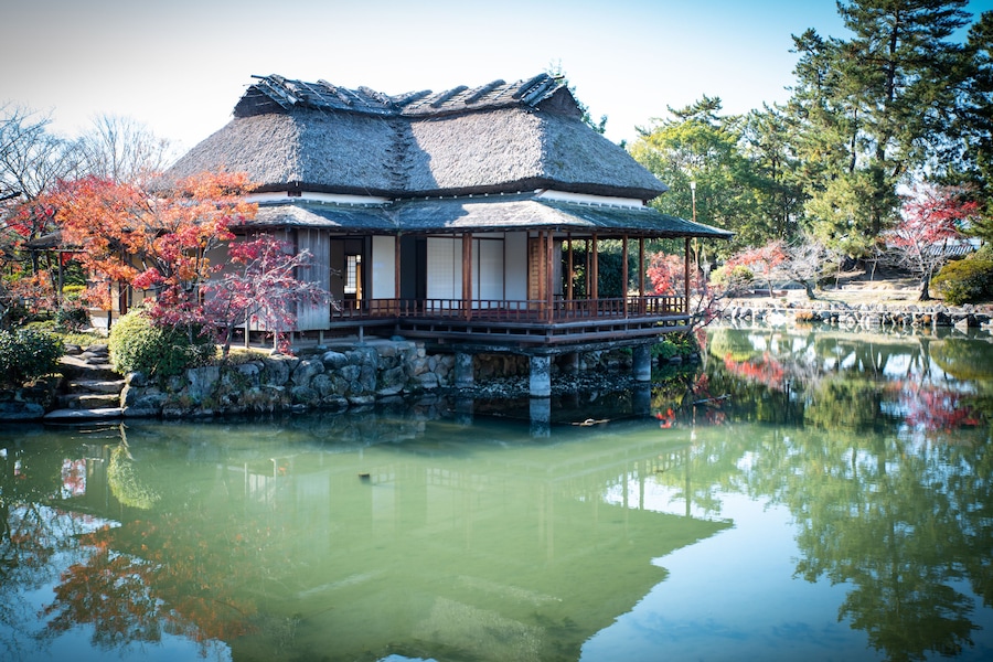 A traditional Japanese building with a thatched roof, surrounded by a pond and autumn-colored trees.
