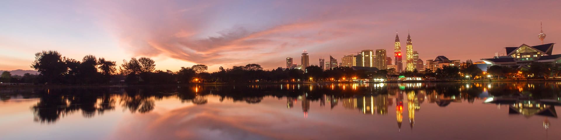 A city skyline at dusk with reflections in a calm body of water, including prominent buildings and a tower.