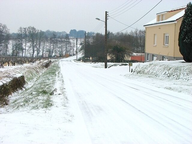 A snow-covered road with a house on the right and bare trees in the background.