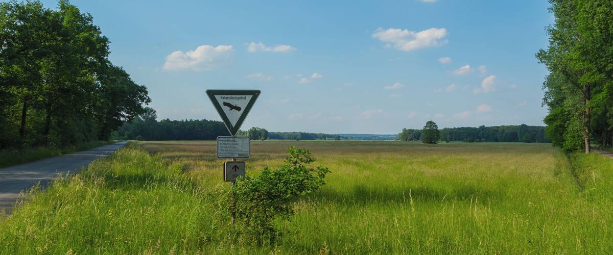 A rural landscape with a nature protection sign, a road, and a grassy field.