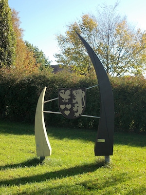 Una escultura de un escudo de caballero con un león y dos aves, ubicada en un parque cubierto de hierba con árboles al fondo.