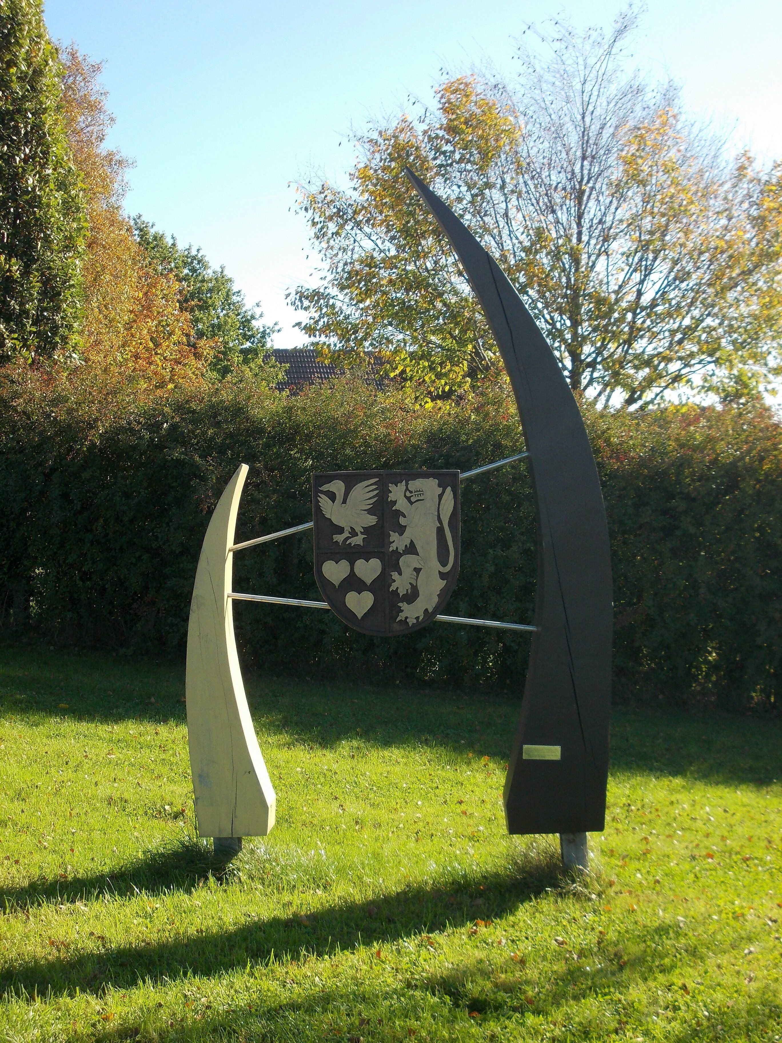 A sculpture of a knight's shield with a lion and two birds, set in a grassy park with trees in the background.