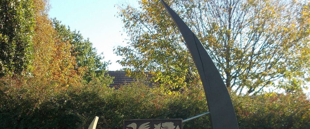 A sculpture of a knight's shield with a lion and two birds, set in a grassy park with trees in the background.