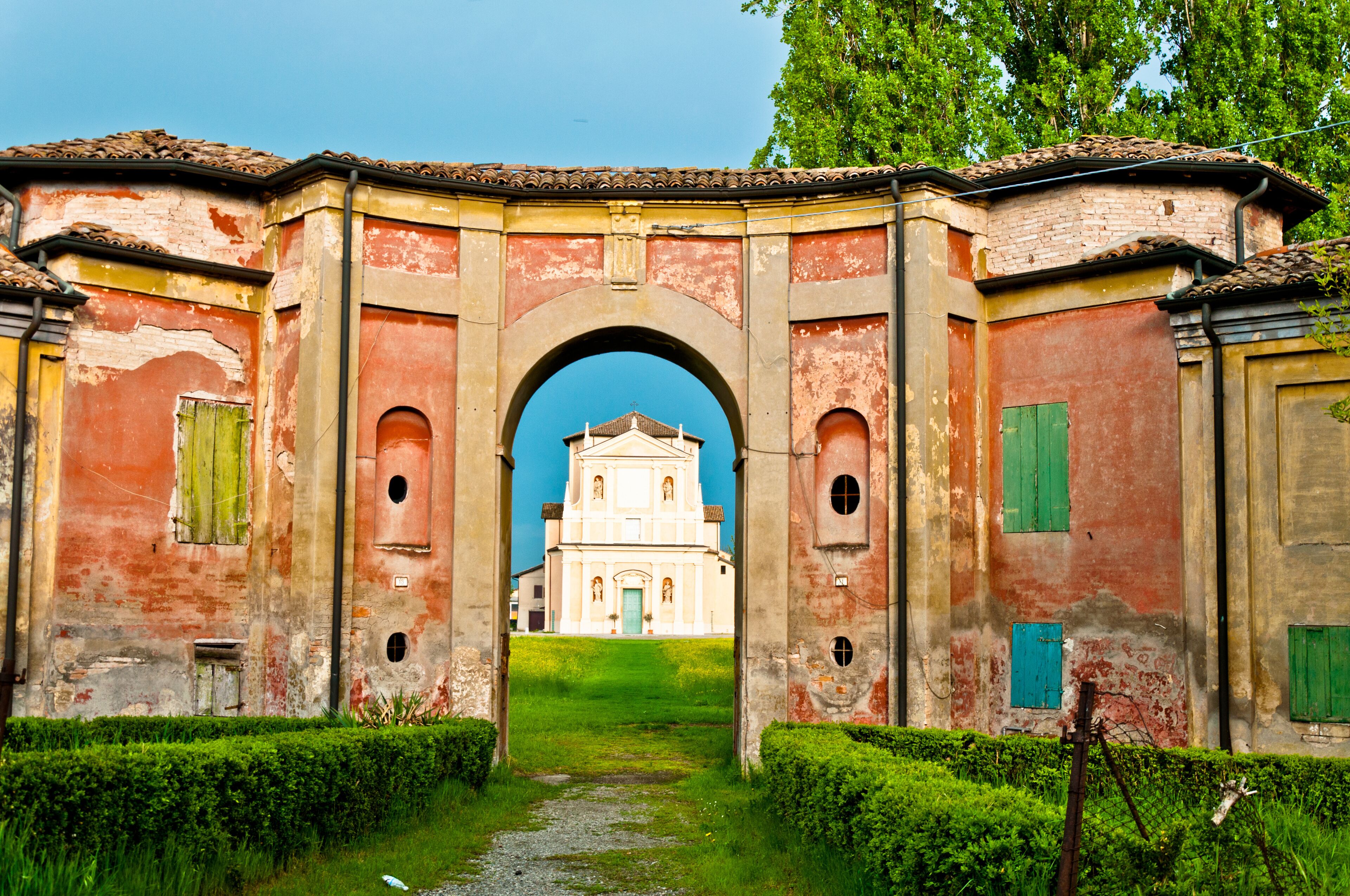 An old, weathered archway leading to a white church with green shutters.