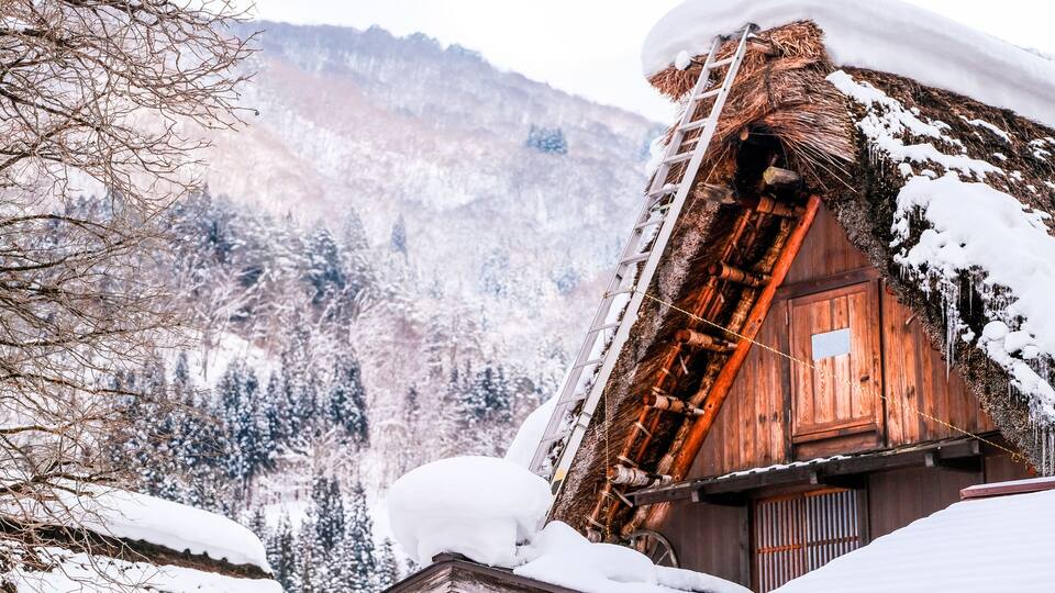 A wooden cabin with a steep roof, surrounded by snow-covered trees and mountains.