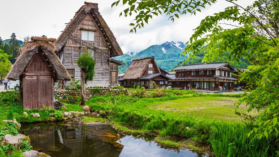 Traditional wooden houses with thatched roofs, a pond with koi fish, and a mountainous backdrop.