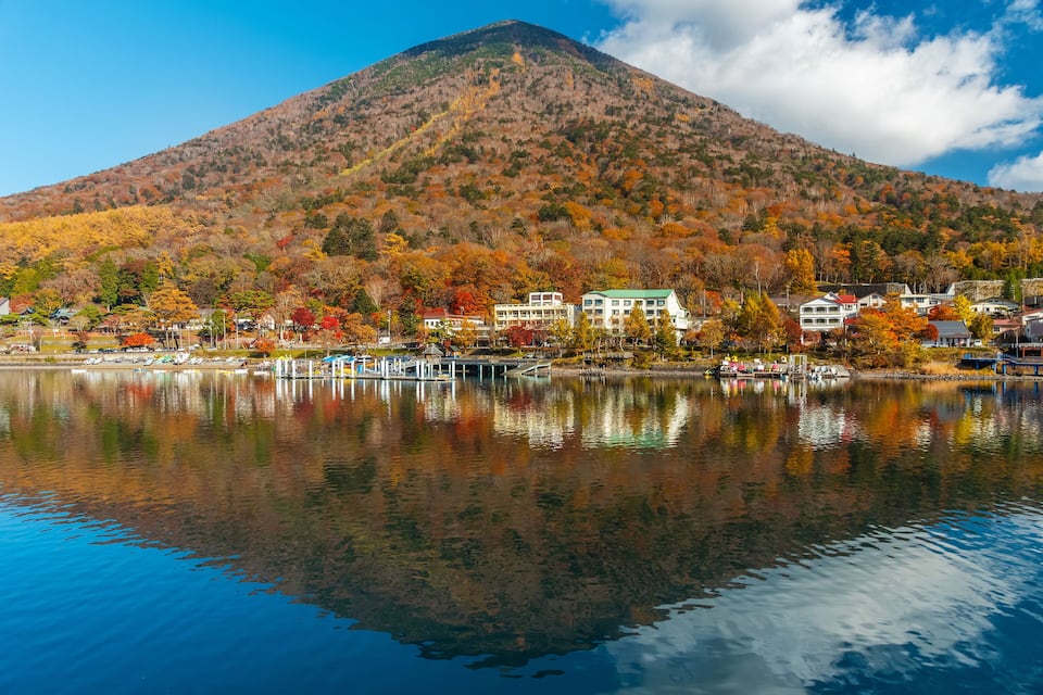 A mountain with autumn foliage, a calm lake reflecting the scenery, and a small town with buildings along the shore.