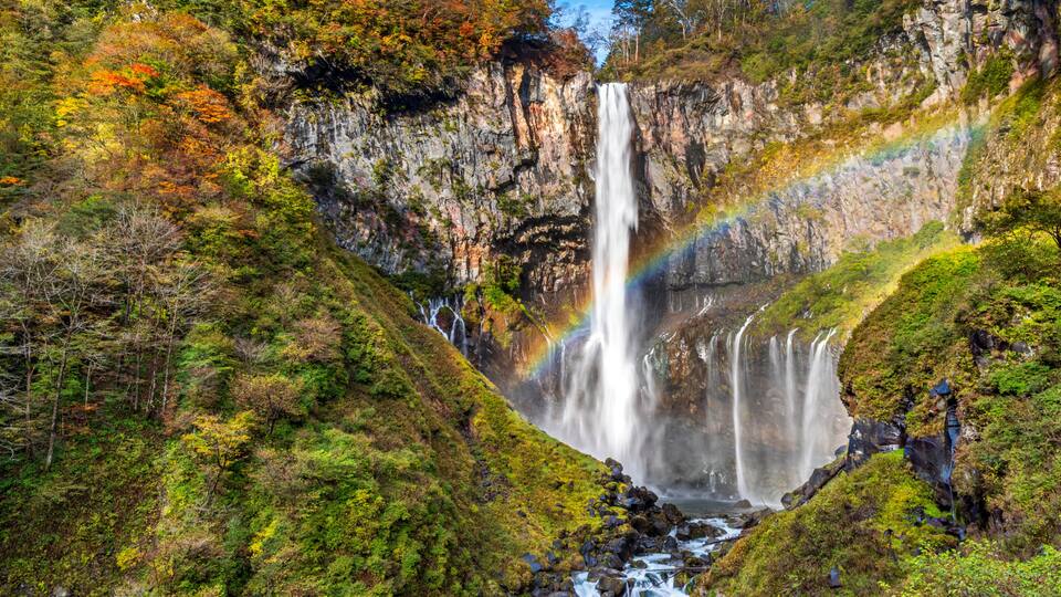 A waterfall with a rainbow, surrounded by lush greenery and rocky cliffs.