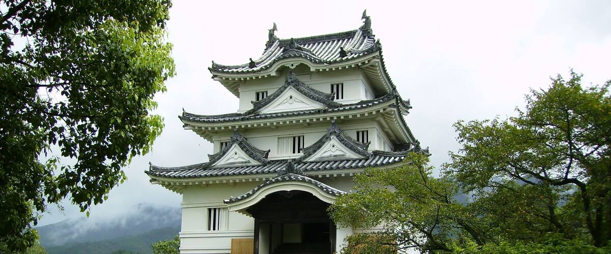 A traditional Japanese castle with multiple tiers and ornate roofs, surrounded by lush greenery and a mountainous backdrop.