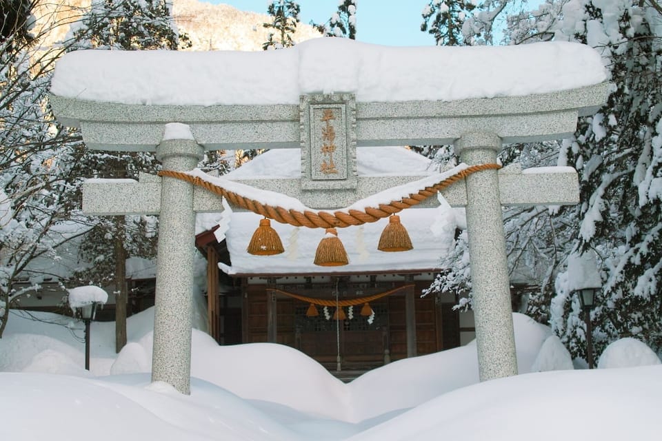 A snow-covered torii gate with a wooden structure behind it.
