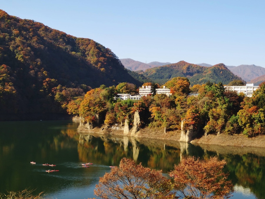 Un lago tranquilo con barcos, rodeado de árboles de colores otoñales y un edificio blanco en una colina.