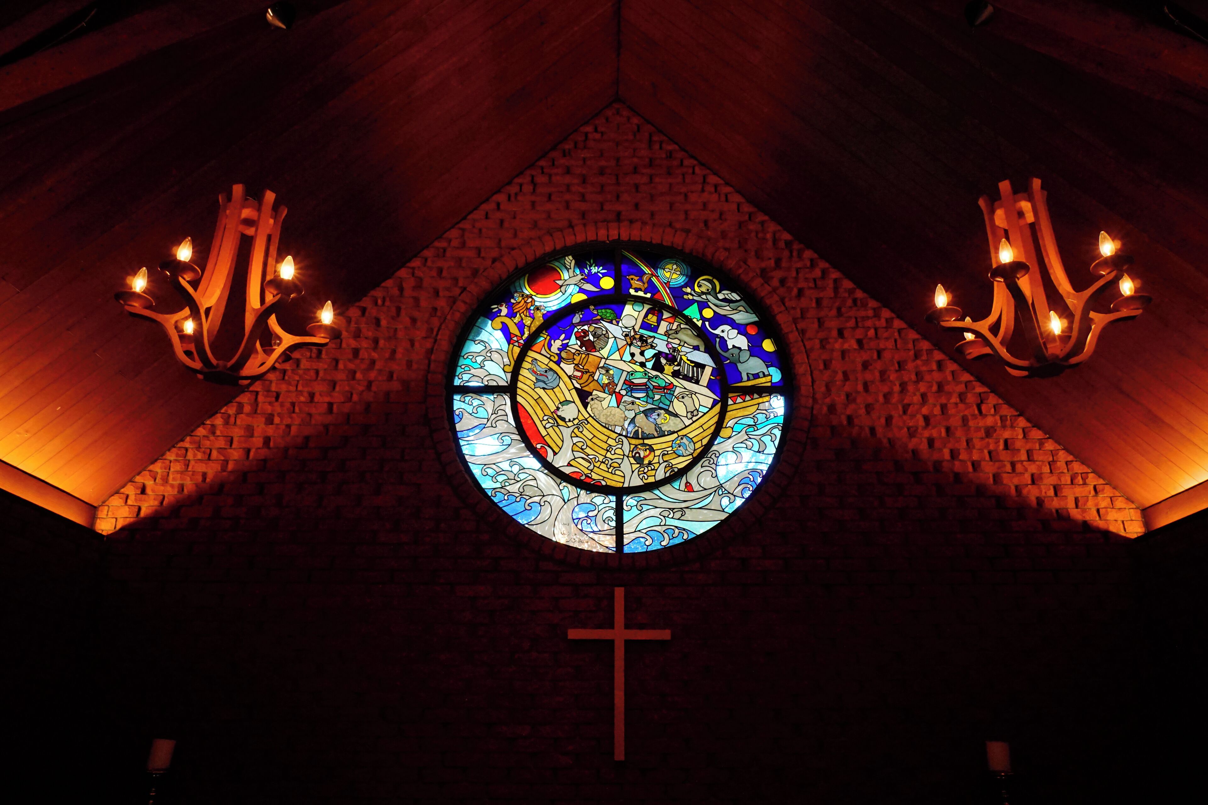 A stained glass window with a cross below it, surrounded by brick walls and two chandeliers.