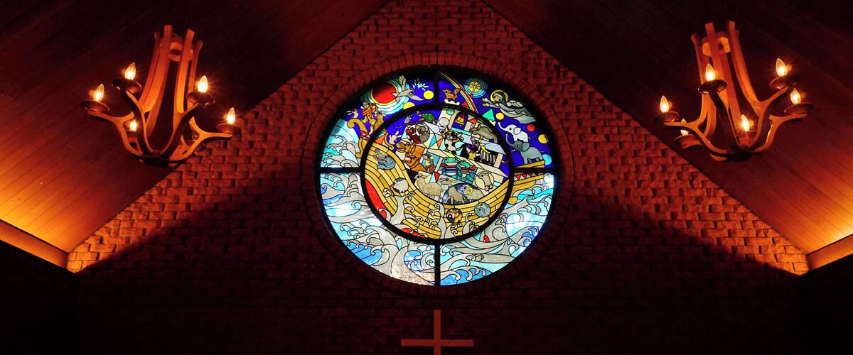 A stained glass window with a cross below it, surrounded by brick walls and two chandeliers.