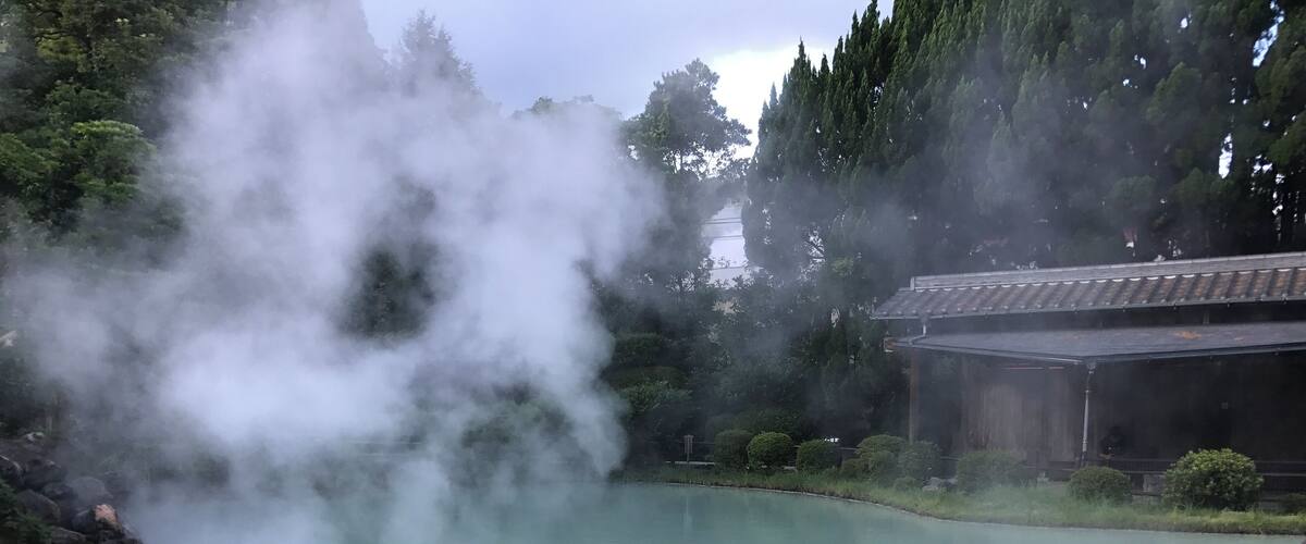 A hot spring with steam rising from the water, surrounded by trees and a traditional building.