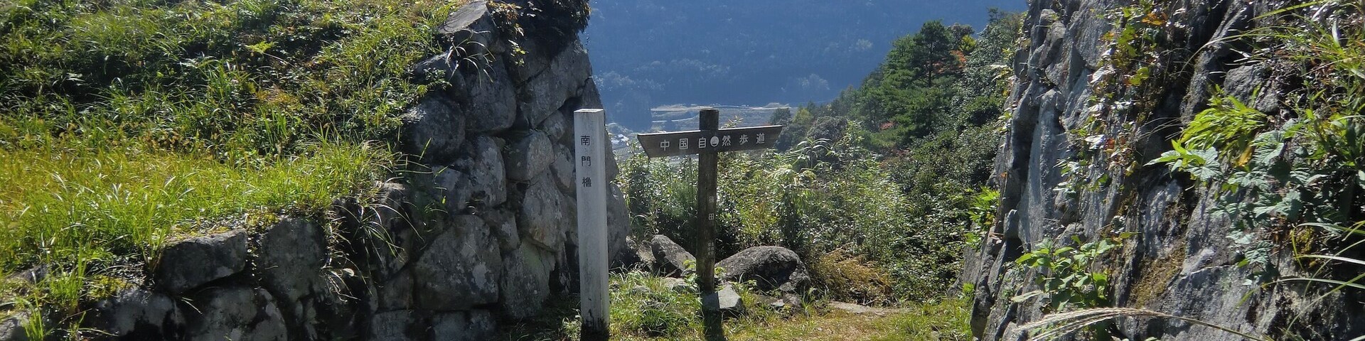 Un sendero de piedra serpentea por un paisaje montañoso con un indicador señalando las direcciones.