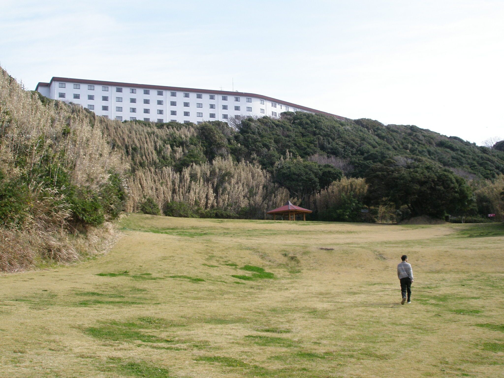 A grassy field with a person walking towards a red-roofed pavilion, a white building in the background, and a hill with dense vegetation.