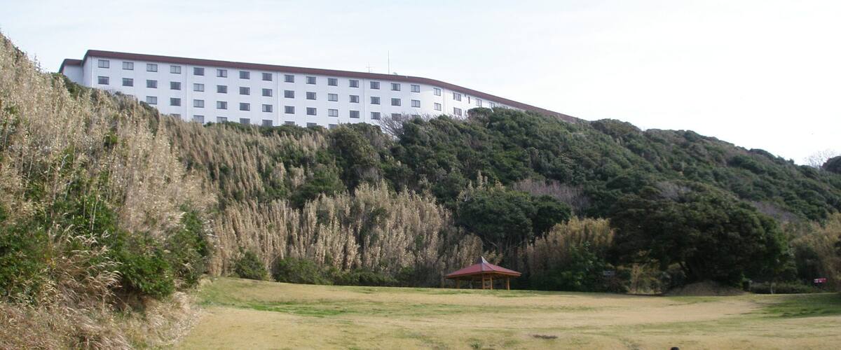 A grassy field with a person walking towards a red-roofed pavilion, a white building in the background, and a hill with dense vegetation.