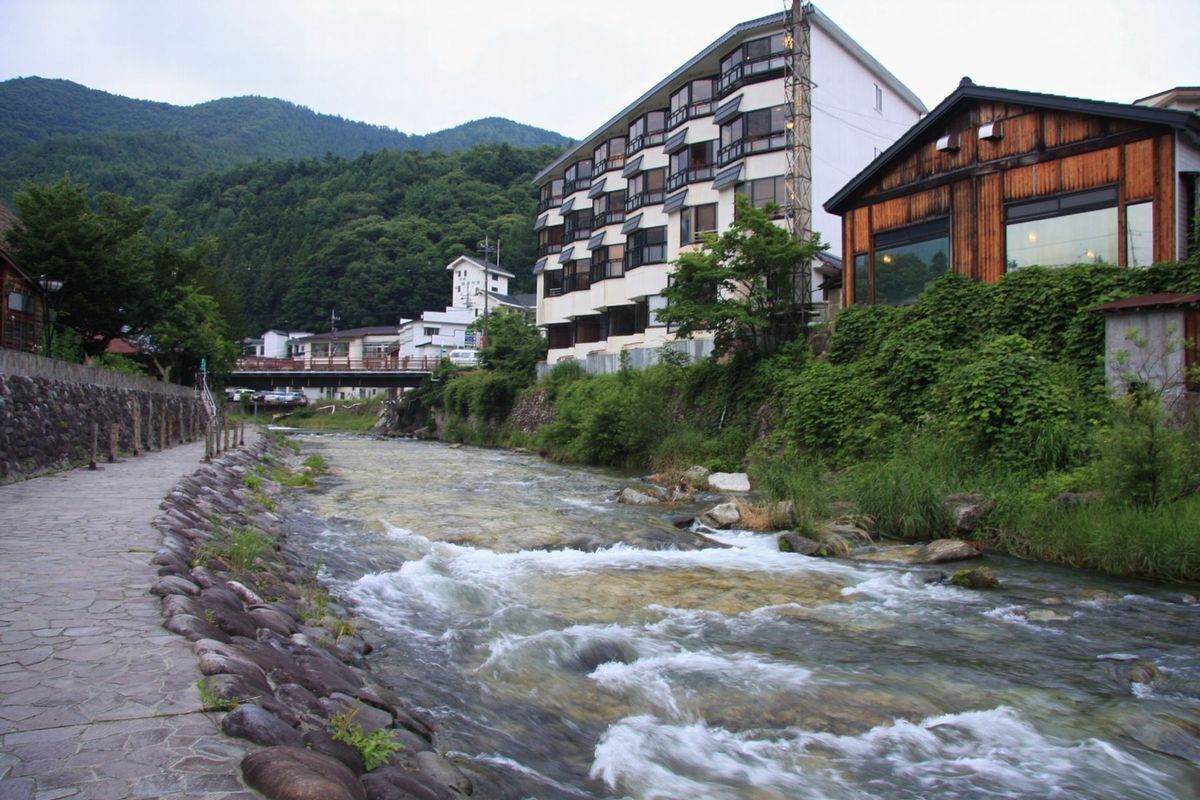 A river flowing through a town with buildings on both sides, surrounded by greenery and mountains.
