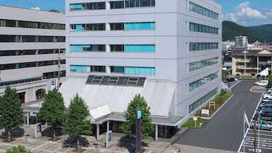 A modern multi-story building with a flat roof and large windows, surrounded by a parking lot and trees.