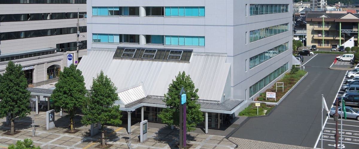 A modern multi-story building with a flat roof and large windows, surrounded by a parking lot and trees.