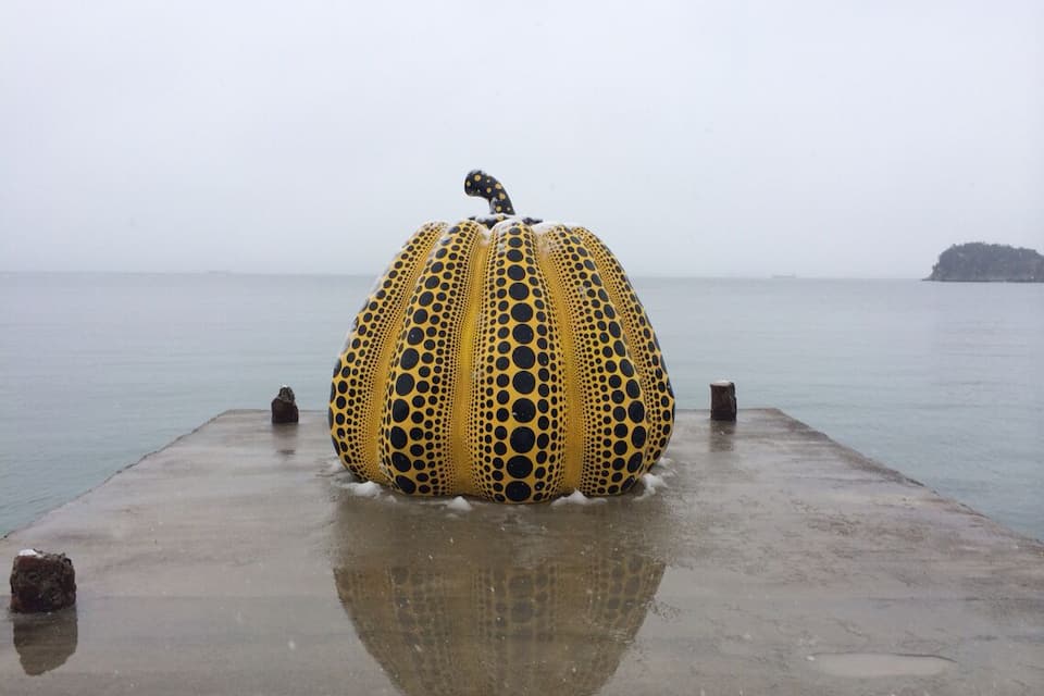 Una gran escultura amarilla y negra con lunares sobre un muelle de hormigón que se adentra en una masa de agua tranquila.