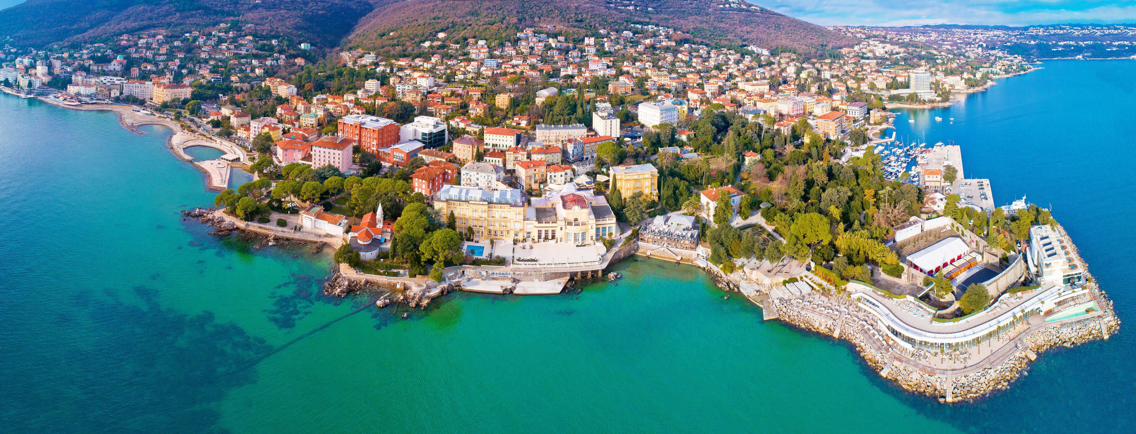 An aerial view of a coastal city with a mix of historic and modern architecture, a marina, and a mountainous backdrop.