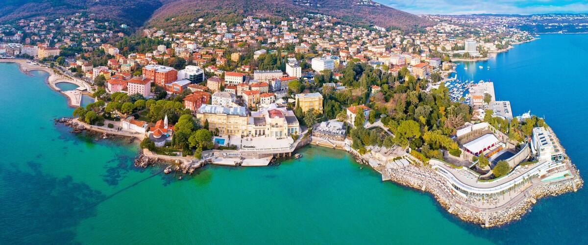An aerial view of a coastal city with a mix of historic and modern architecture, a marina, and a mountainous backdrop.