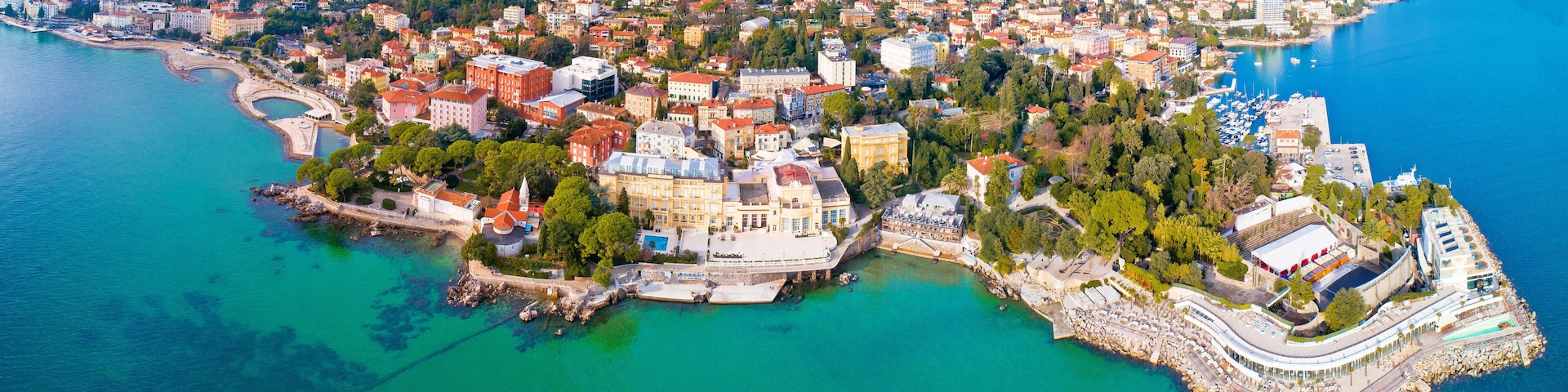 An aerial view of a coastal city with a mix of historic and modern architecture, a marina, and a mountainous backdrop.
