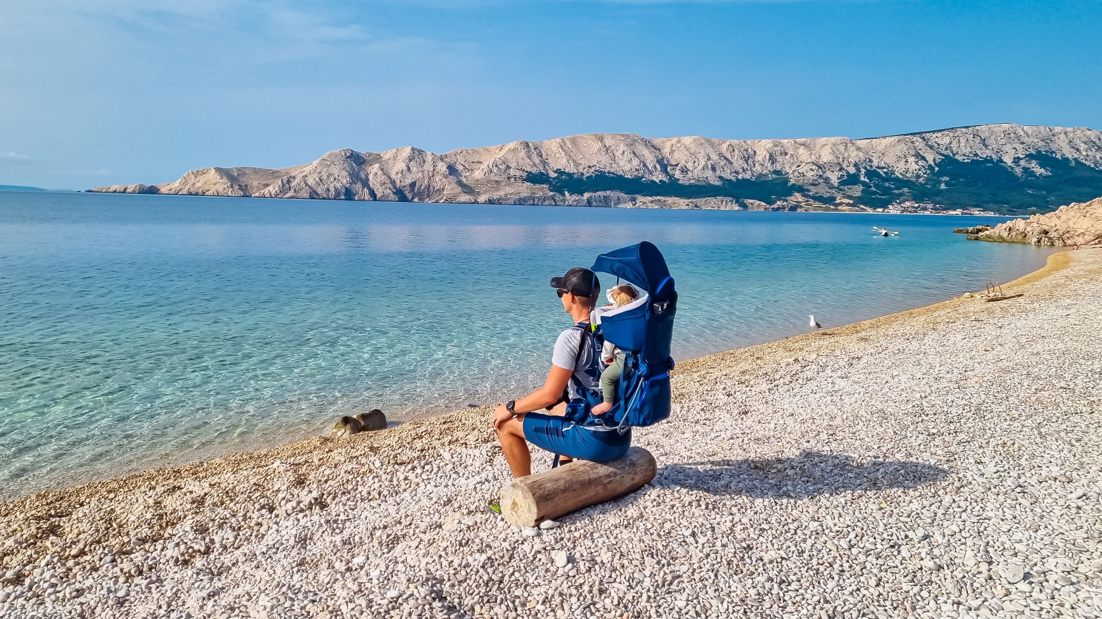 A person sitting on a log by a pebble beach with a child in a blue carrier, overlooking a calm sea and rocky hills.