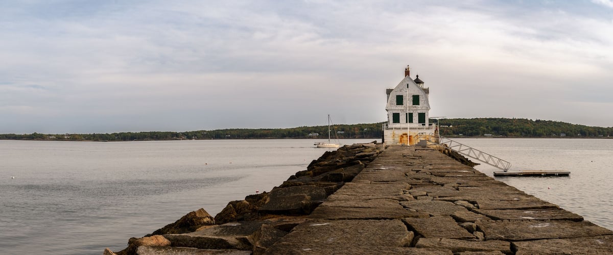 A stone pier leading to a white lighthouse with a green door, overlooking calm waters and a forested island.