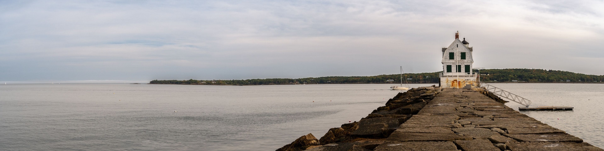 A stone pier leading to a white lighthouse with a green door, overlooking calm waters and a forested island.